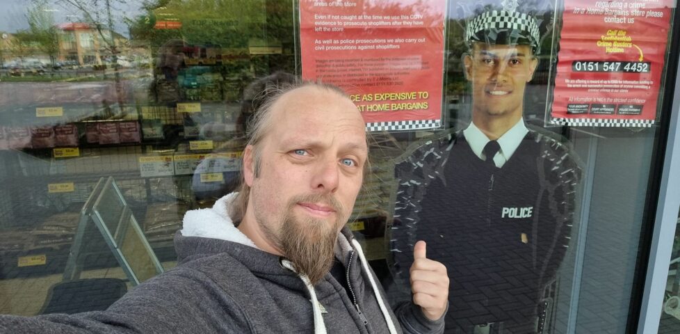 Dan stands outside a floor-to-ceiling shop window within which is a cardboard cut-out of a smiling police officer.