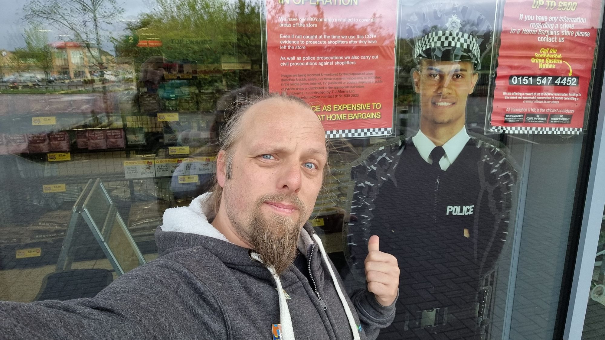 Full image of Dan stands outside a floor-to-ceiling shop window within which is a cardboard cut-out of a smiling police officer.