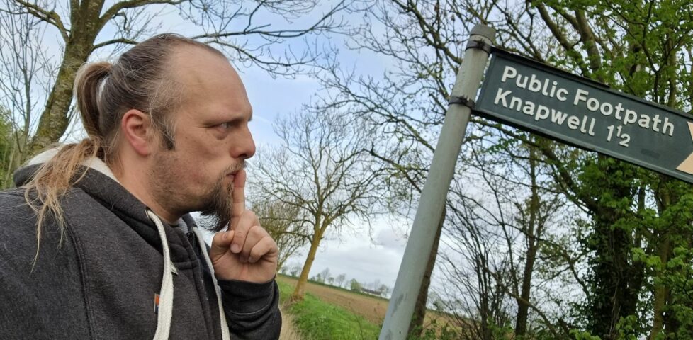 Dan, his finger to his lips as-if in thought, looks at a finger post sign that indicates a public footpath to Knapwell (1ยฝ miles).