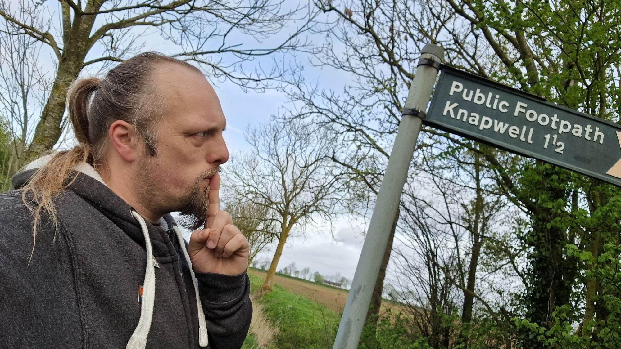 Full image of Dan, his finger to his lips as-if in thought, looks at a finger post sign that indicates a public footpath to Knapwell (1½ miles).