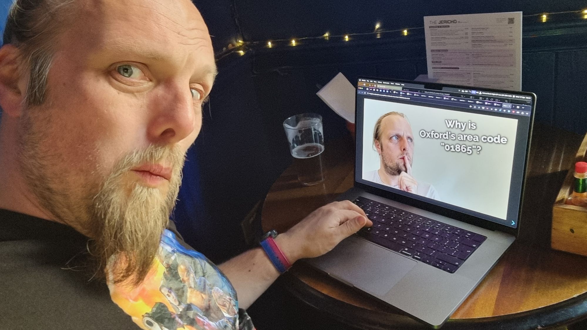 Dan, a white man with a goatee-style beard, wearing a black t-shirt and a bi pride watch strap, sits at a bar table with a half-empty (half-full?) pint of beer and a laptop. He looks mildly concerned. The laptop screen shows Dan's face, deep in thought, alongside the words "Why is Oxford's area code '01865'?"