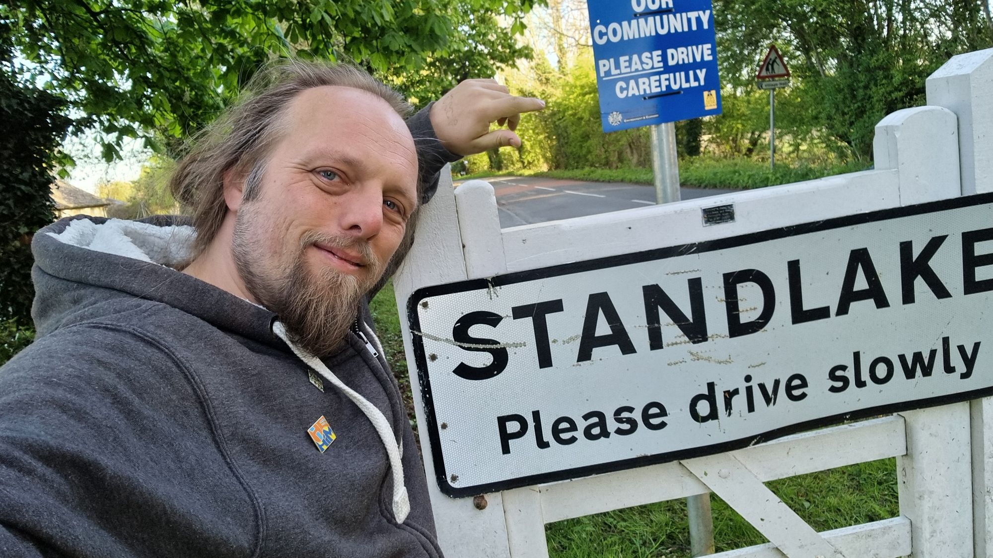 Full image of Dan, wearing a grey hoodie, stands by a sign welcoming visitors to the village of Standlake.