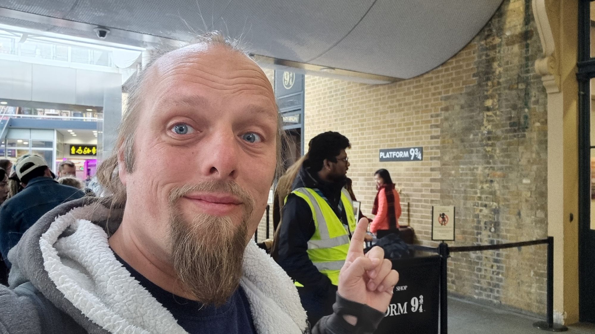 Full image of Dan, a queue of tourists behind him, points over his shoulder beyond them to a sign on a brick wall, "Platform 9¾".