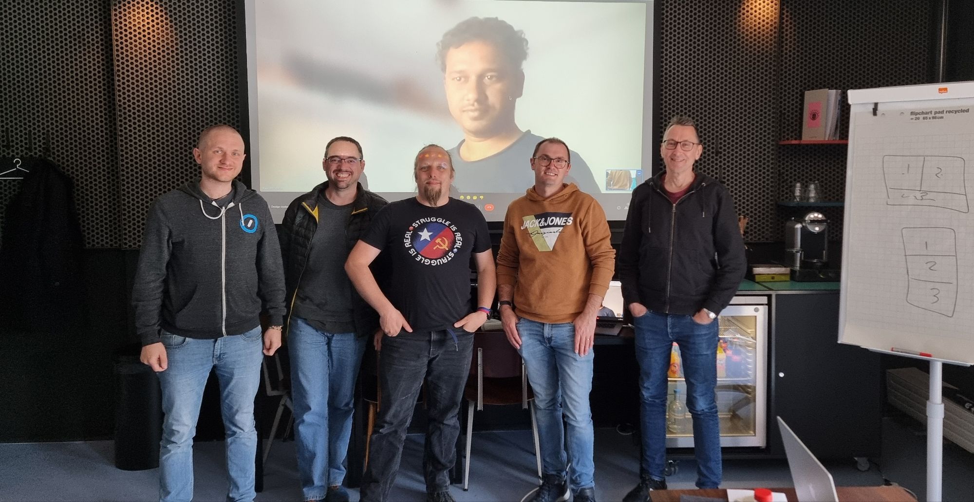 Full image of Dan stands with four other men in front of a projector screen with a fifth man on it.