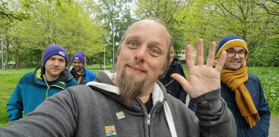 Dan waves to the camera; several other geeky folks walk along behind him in a leafy green city space.