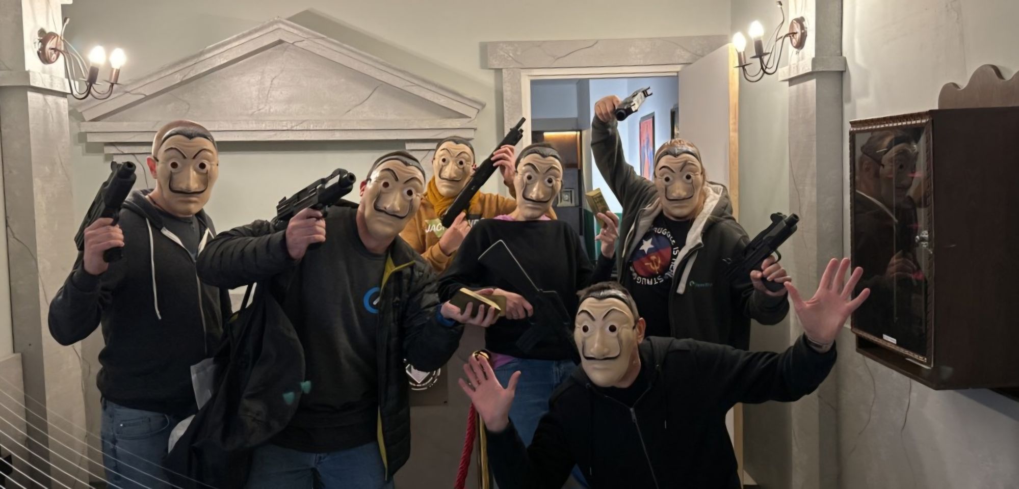 Full image of A group of people dressed as robbers (with masks covering their faces and automatic weapons in their hands) pose menacingly in the lobby of a vault.