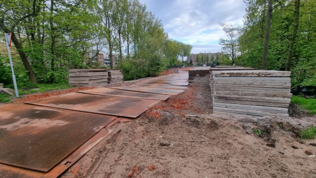 A torn-up footpath adapted for use by construction vehicles, going over a small bridge at the entrance to a park.