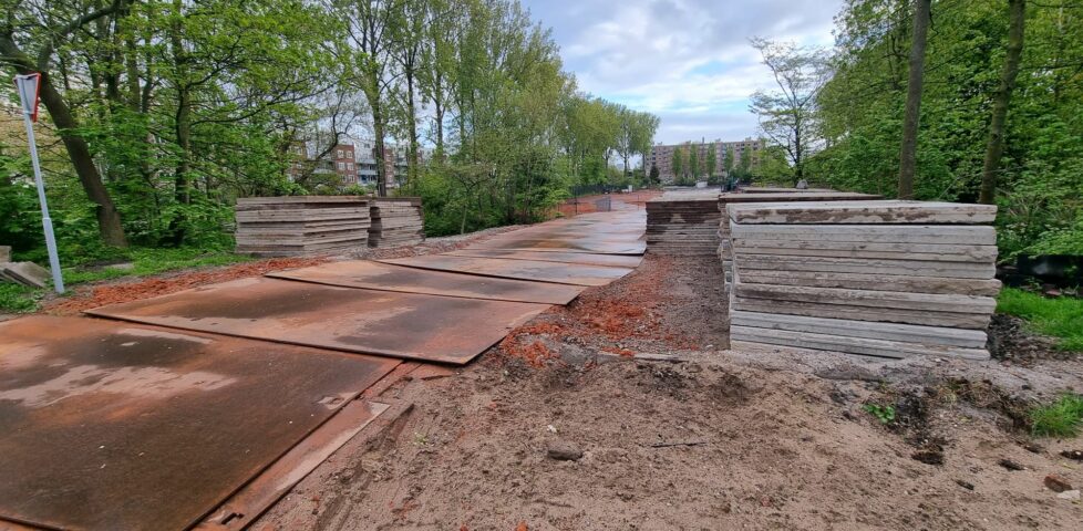 A torn-up footpath adapted for use by construction vehicles, going over a small bridge at the entrance to a park.