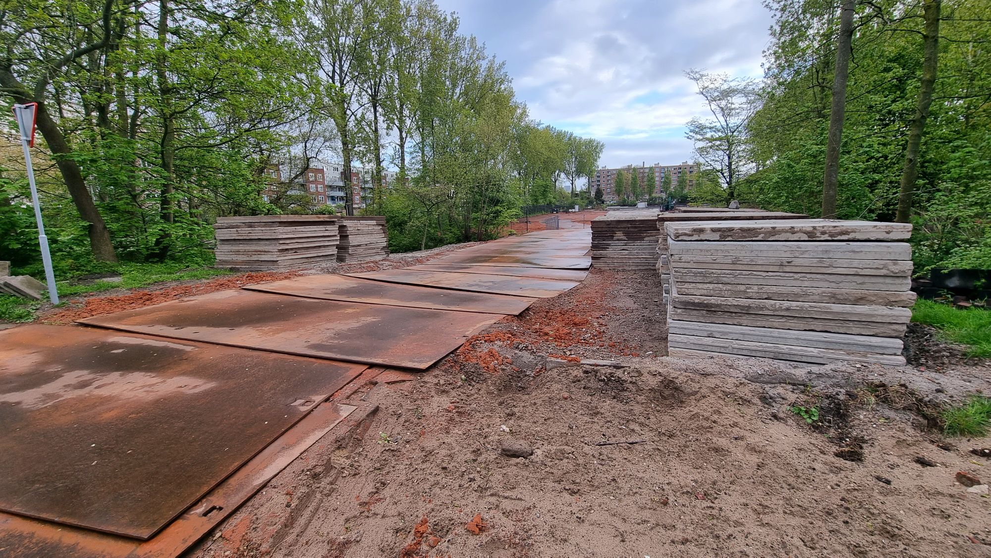 Full image of A torn-up footpath adapted for use by construction vehicles, going over a small bridge at the entrance to a park.