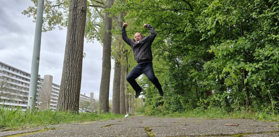 On a tree-lined canalside footpath, Dan leaps theatrically into the air as if in incredible excitement.