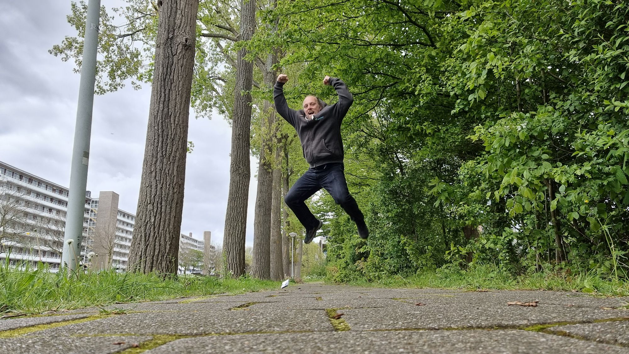 Full image of On a tree-lined canalside footpath, Dan leaps theatrically into the air as if in incredible excitement.