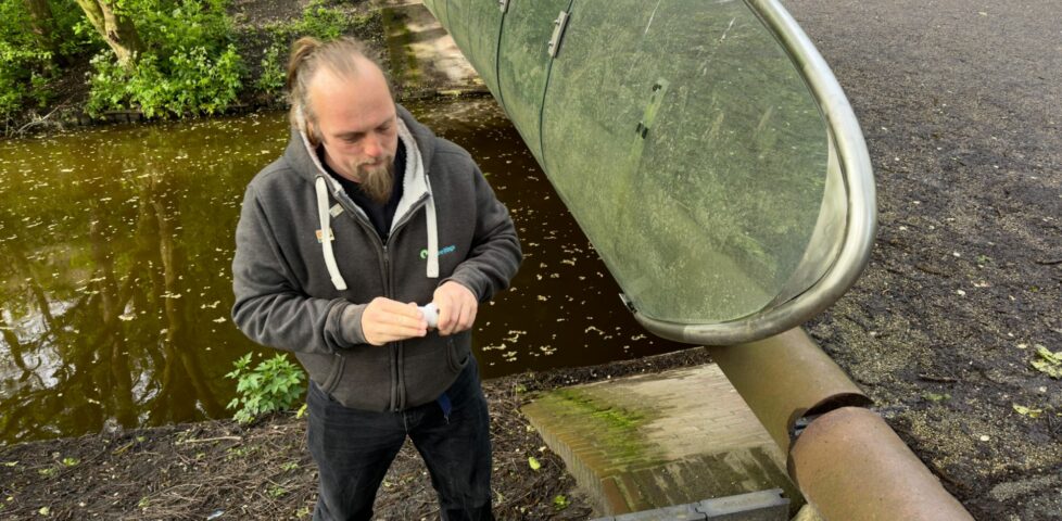 Dan on the bank of a water course, beneath a bridge, unrolls a logbook to sign his name.
