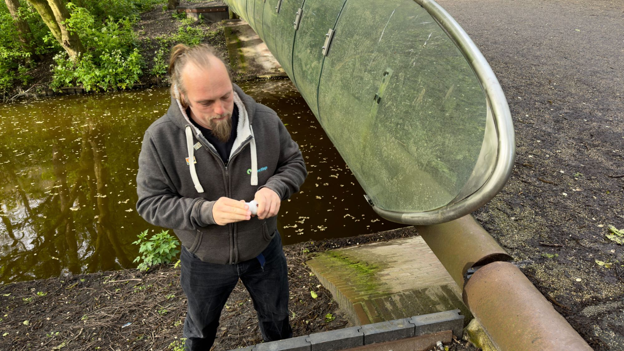 Full image of Dan on the bank of a water course, beneath a bridge, unrolls a logbook to sign his name.