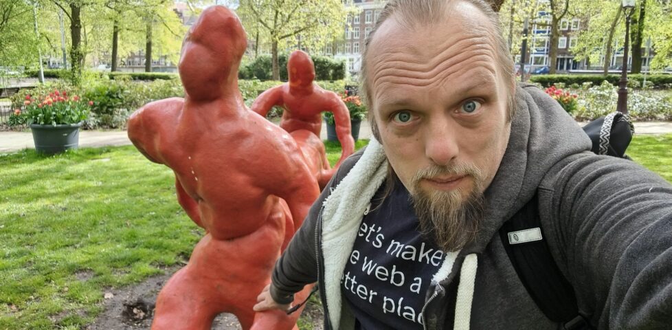 Dan holds the hand of a statue, as if guiding it, as it "carries" a log through a park.
