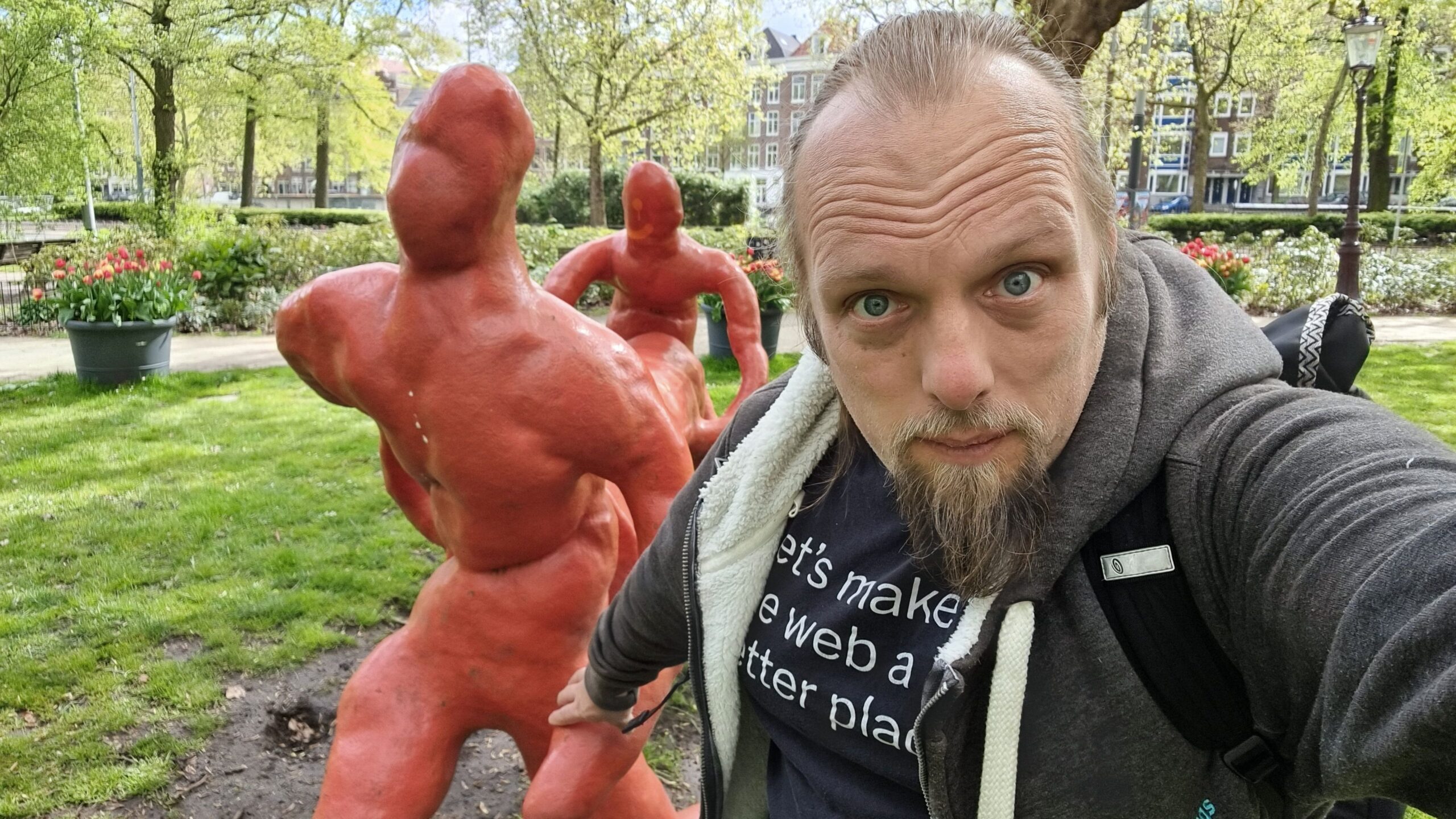 Full image of Dan holds the hand of a statue, as if guiding it, as it "carries" a log through a park.