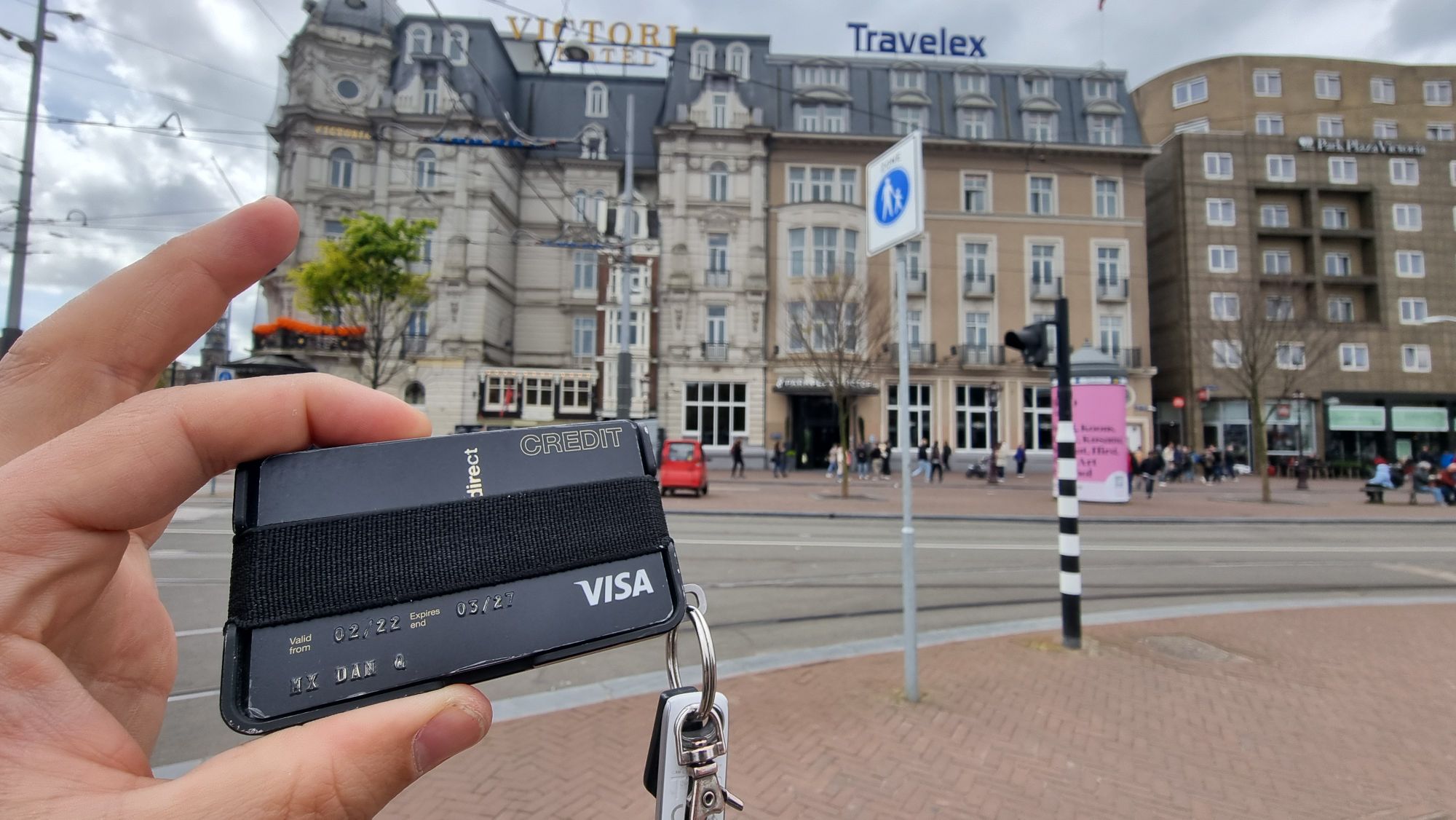 Full image of Dans fingers hold up a minimalist carbon fibre wallet in front of the Victoria Hotel in central Amsterdam. His credit card is visible showing his name but little else.