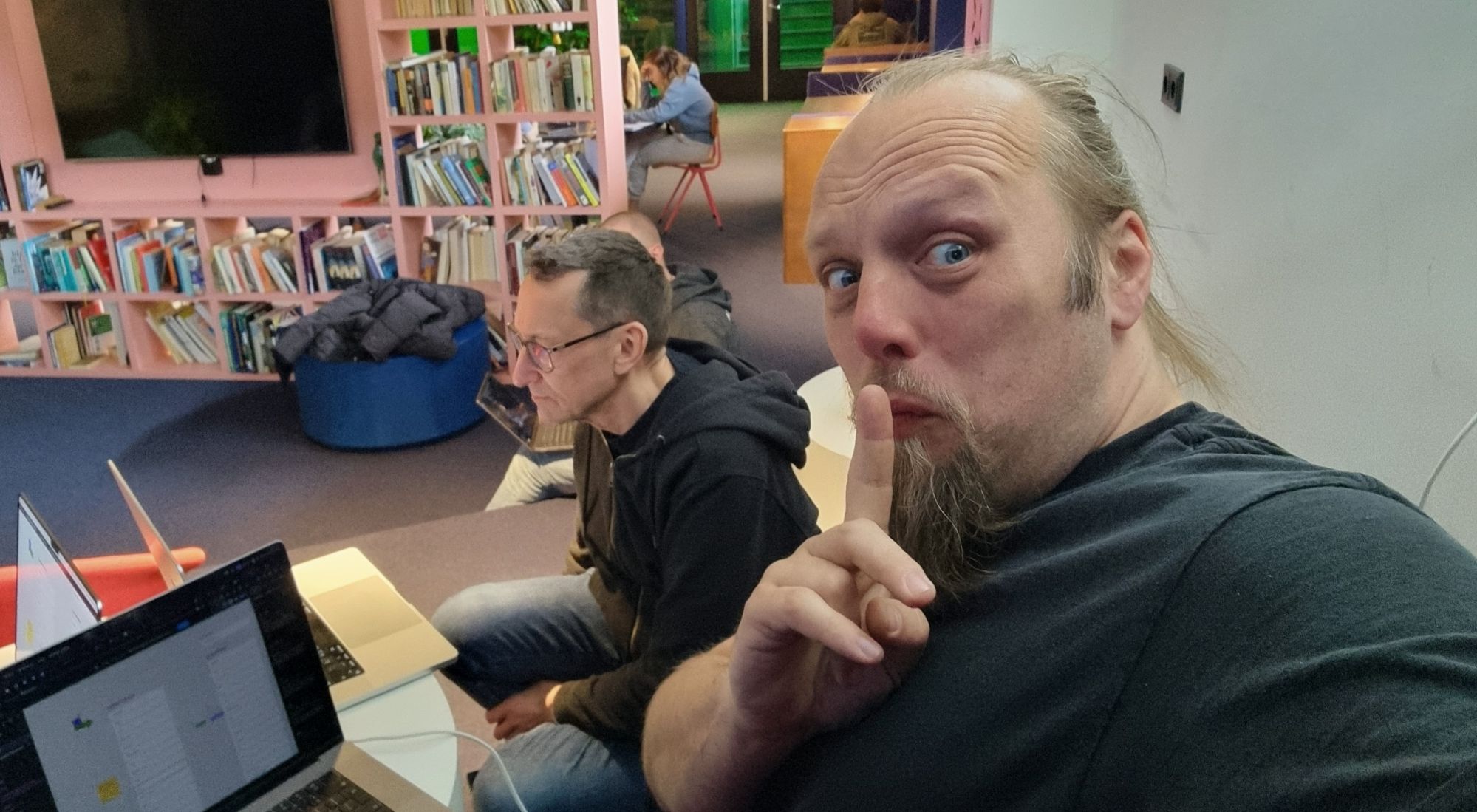 Full image of Dan, his finger to his lips and his laptop on his knees, makes a "shush" action. A coworker can be seen working behind him.
