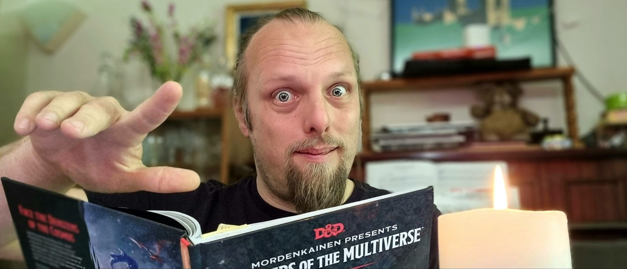 Full image of Dan gestures with a wave as he peers into the camera over the top of a copy of Monsters of the Multiverse, by candlelight.