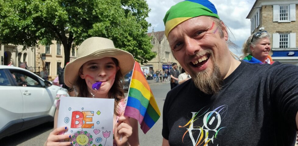 A tweeny girl and a 40-something man with rainbows painted on their faces wave flags in a Pride parade. The child has coloured-in a poster saying "be kind".