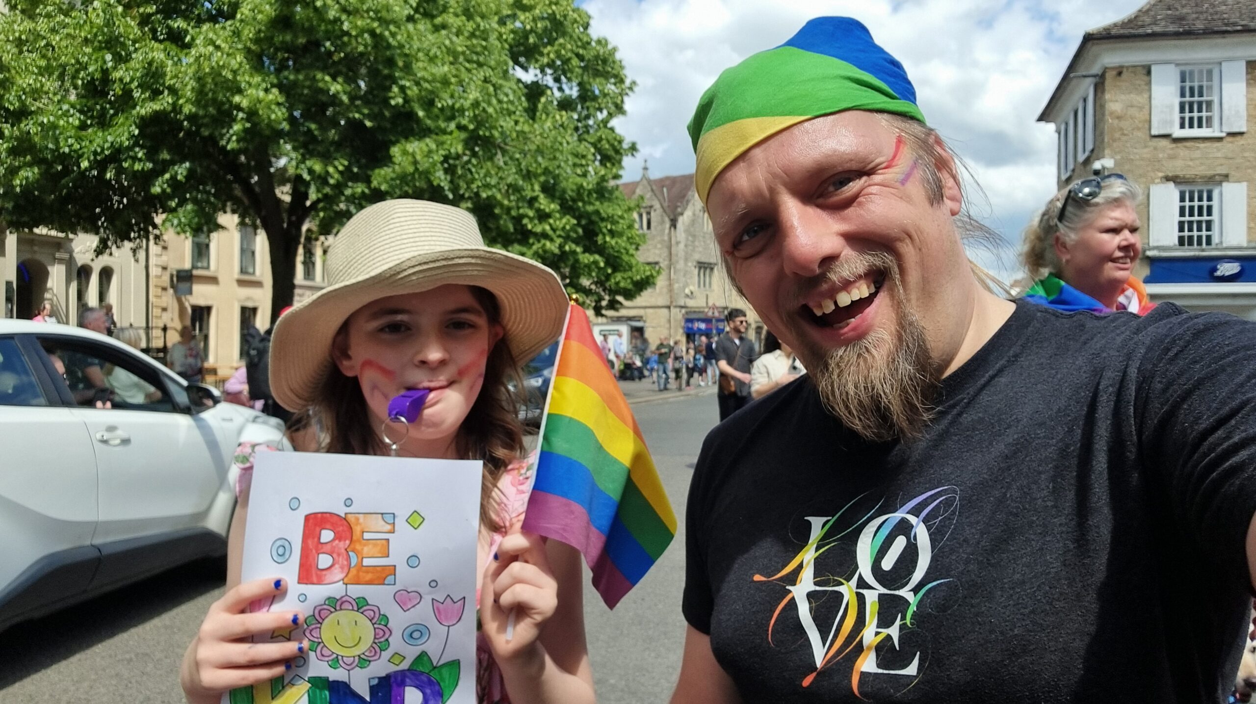 Full image of A tweeny girl and a 40-something man with rainbows painted on their faces wave flags in a Pride parade. The child has coloured-in a poster saying "be kind".