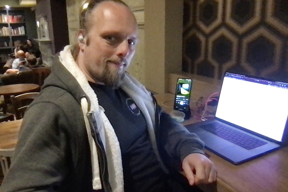 Full image of Dan sits at his laptop in a cafe; his phone is playing music to his earbuds.