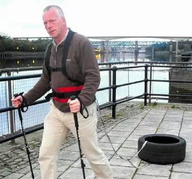 A middle-aged man carrying walking poles on an urban riverbank drags a car tyre that's chained to his waist.