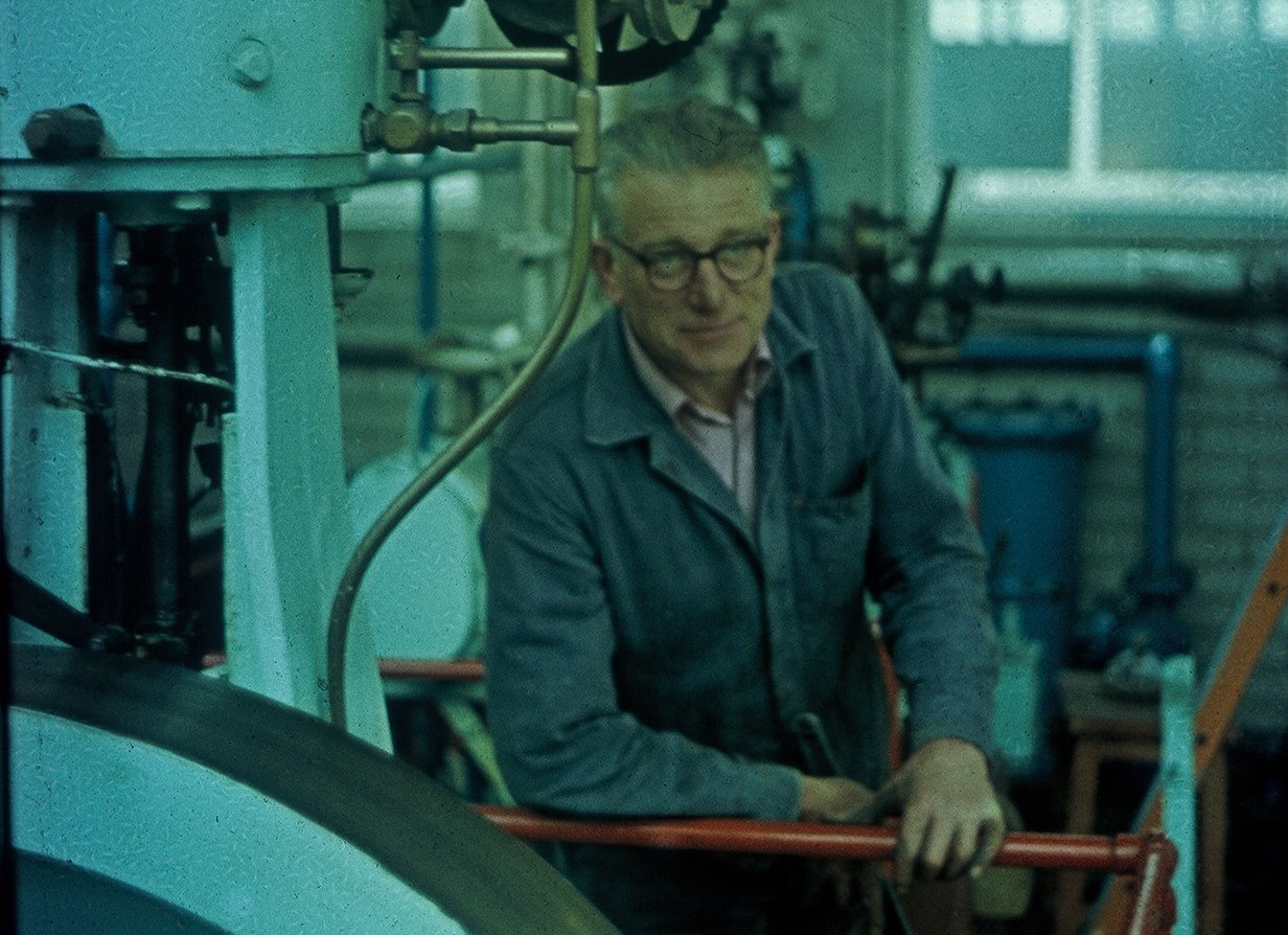 Full image of A grey-haired white man wearing spectacles and a boiler suit leans comfortably on a railing alongside industrial machinery.