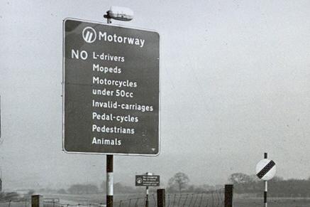 Monochrome photograph showing a sign at the entrance to the Preston By-pass, reading: 'Motorway. NO L-drivers, mopeds, motorcycles under 50cc., invalid-carriages, pedal-cycles, pedestrians, animals'.