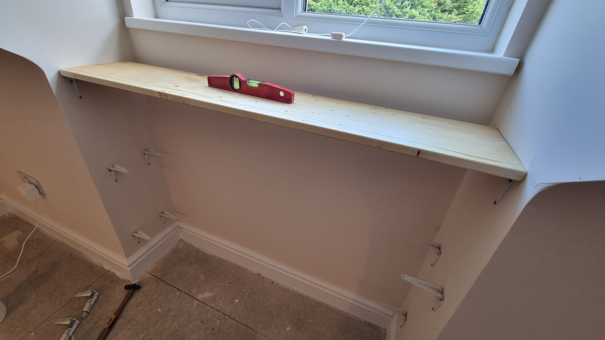 Full image of A spirit level on an unfinished shelf, under a window and beneath an uncarpeted floor.