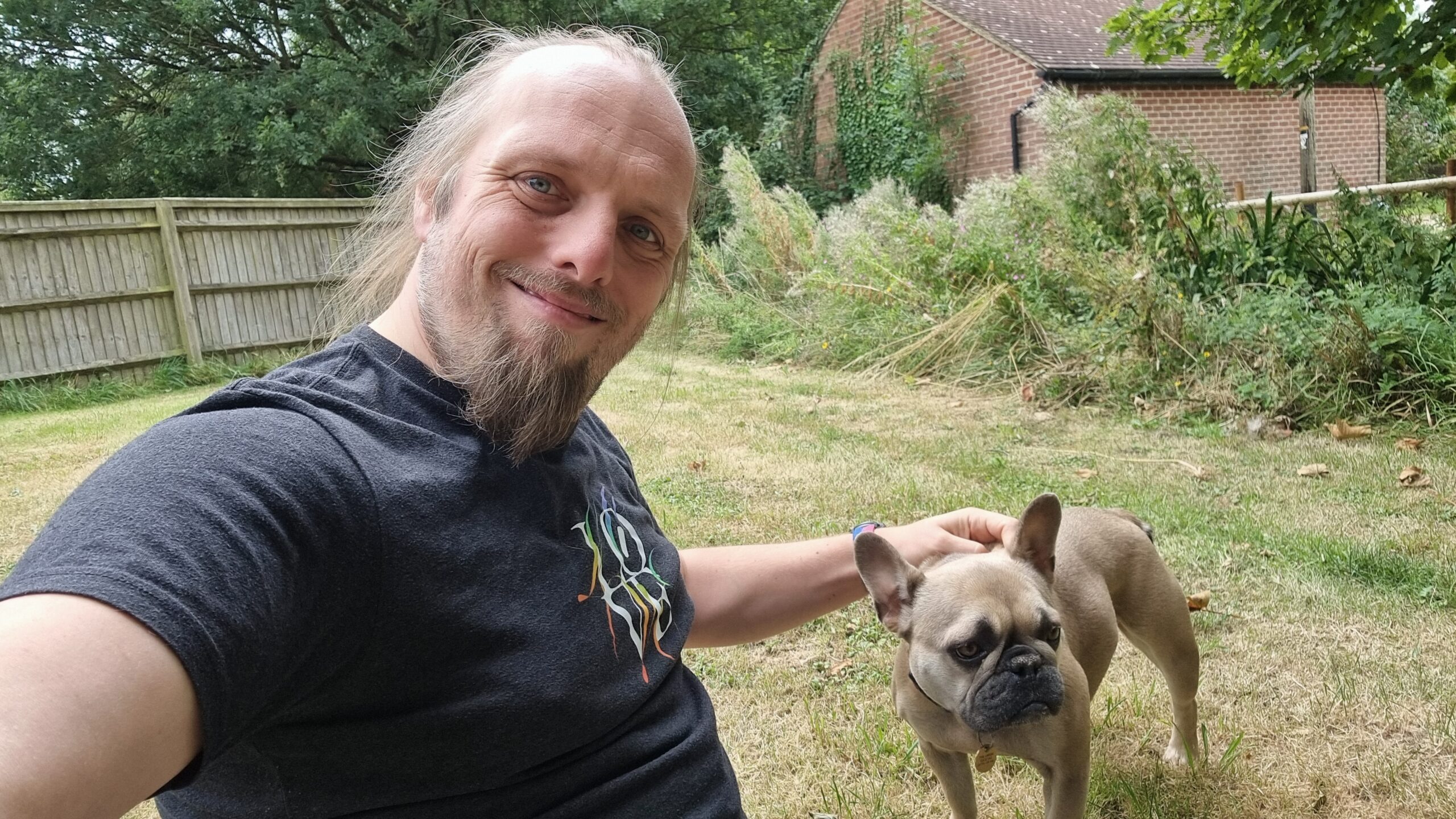 Full image of Dan sits on the grass in a garden, with his French Bulldog.