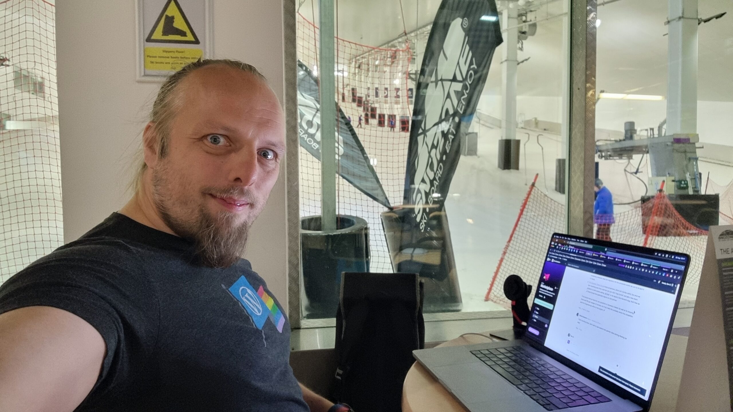 Full image of Dan sits at his laptop in a cafe; ski lifts can be seen in the background.