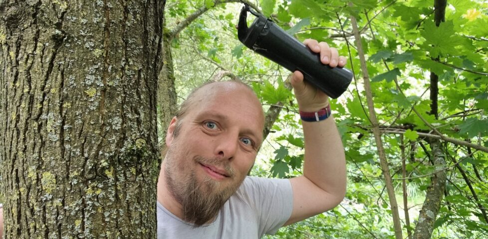 Dan, up a tree, holds a geocache secured high up the trunk.