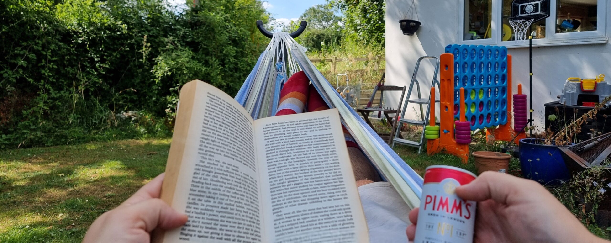 First-person view down a hammock in a rural garden. The hammock's occupier (Dan) is holding a novel and a can of premixed Pimms & lemonade.
