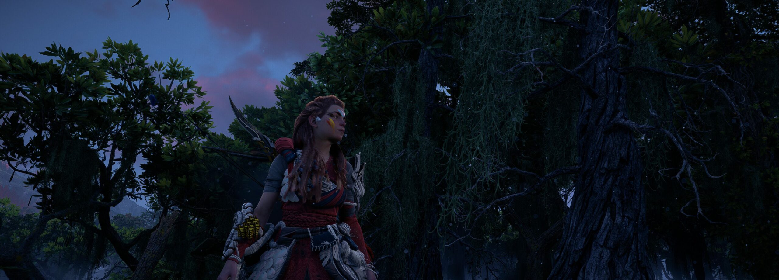 Full image of Aloy, the protagonist of the Horizon games, wearing Mark of Pride face paint and red-stained Quen Deadeye armour, stands at sunset in a jungle environment.