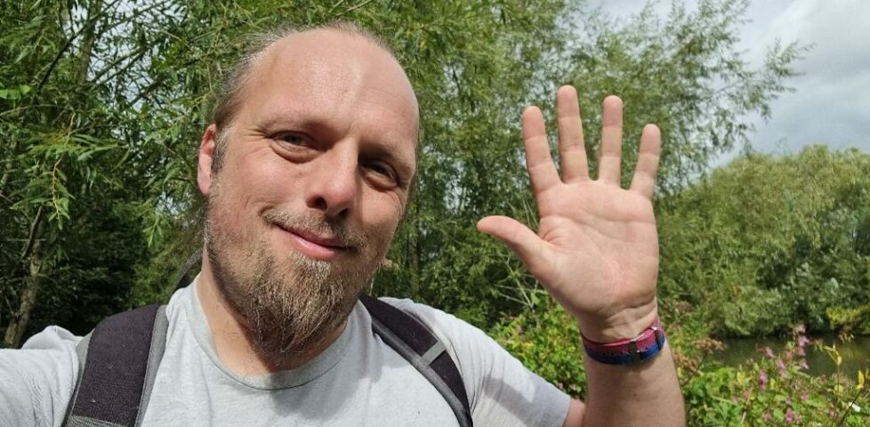 Dan, on an overgrown riverside footpath, waves to the camera.