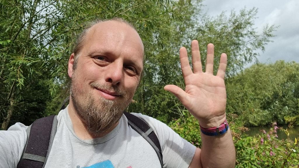 Full image of Dan, on an overgrown riverside footpath, waves to the camera.