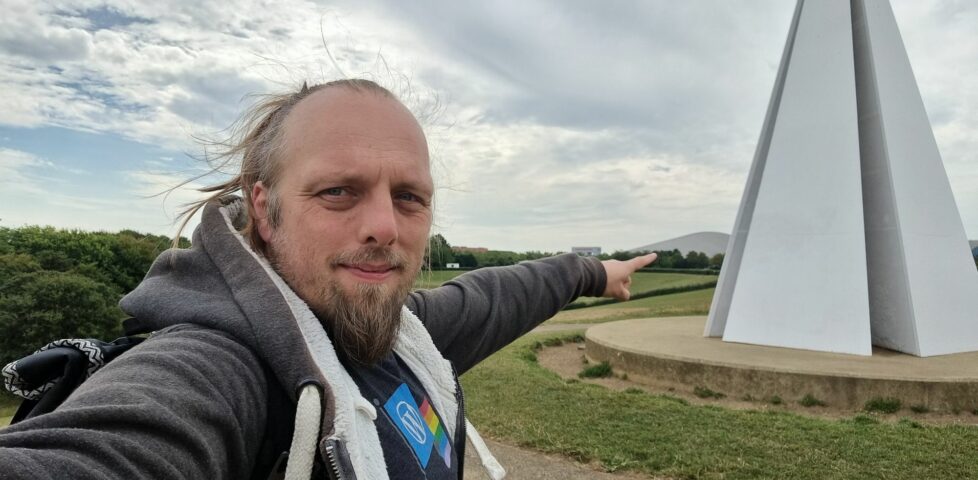 Dan, near a white pyramid-shaped sculpture, points at a distant hill-shaped building.