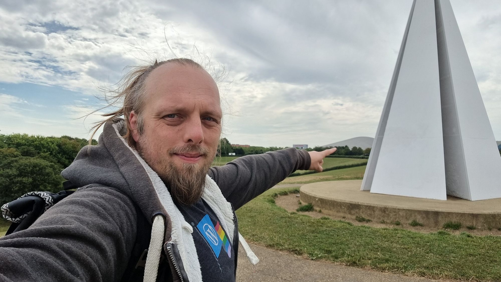 Full image of Dan, near a white pyramid-shaped sculpture, points at a distant hill-shaped building.