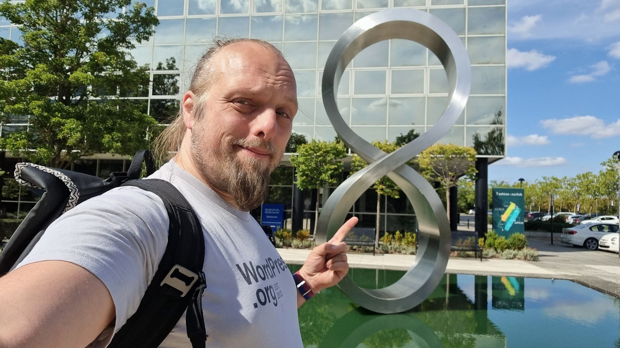 Full image of Dan poses for a selfie in front of a figure-8/moebius strip sculpture, on a sunny day.