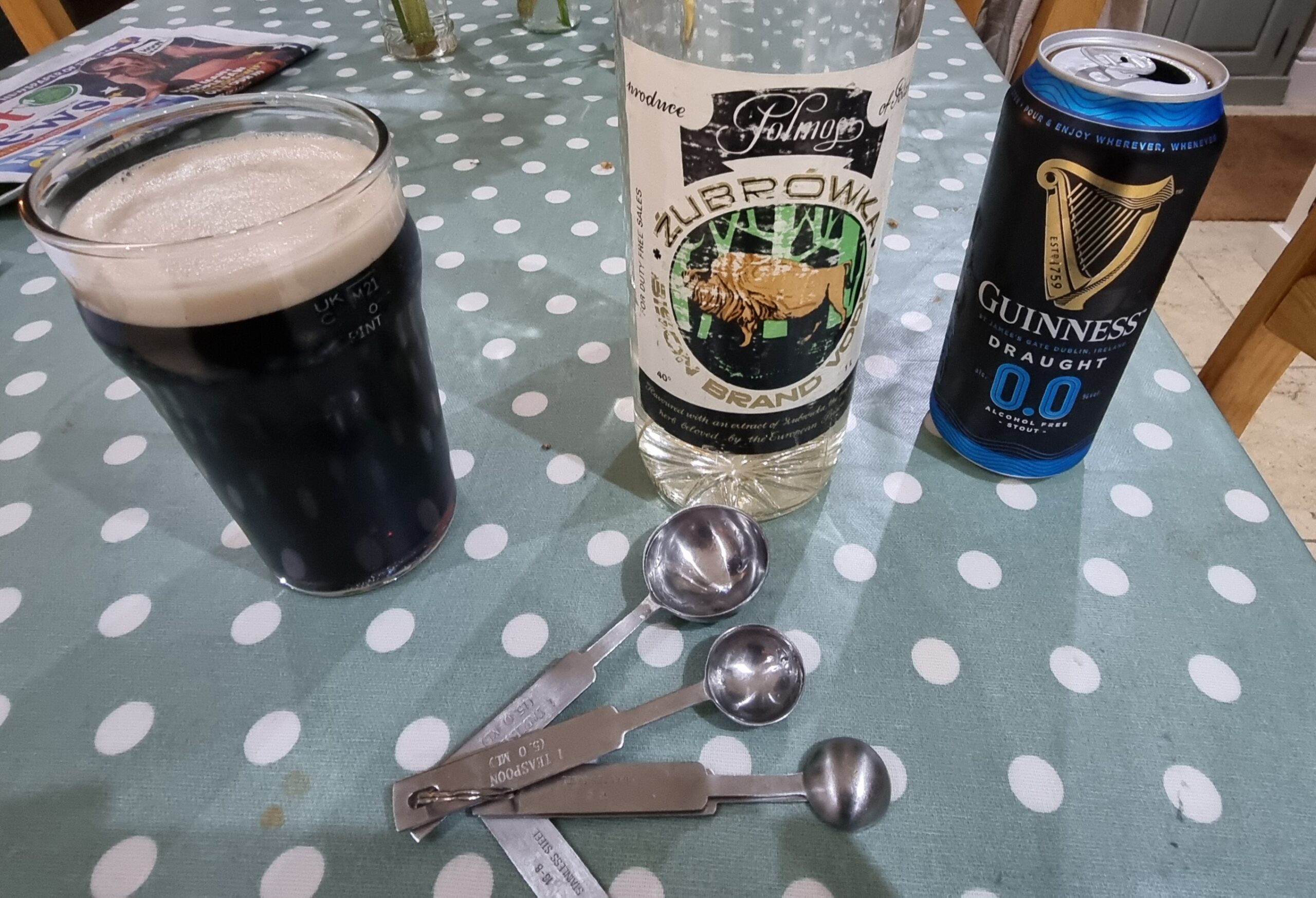 Full image of Pint glass having been filled with a carefully measured mixture of 0%ABV Guinness and vodka, with measuring spoons alongside.