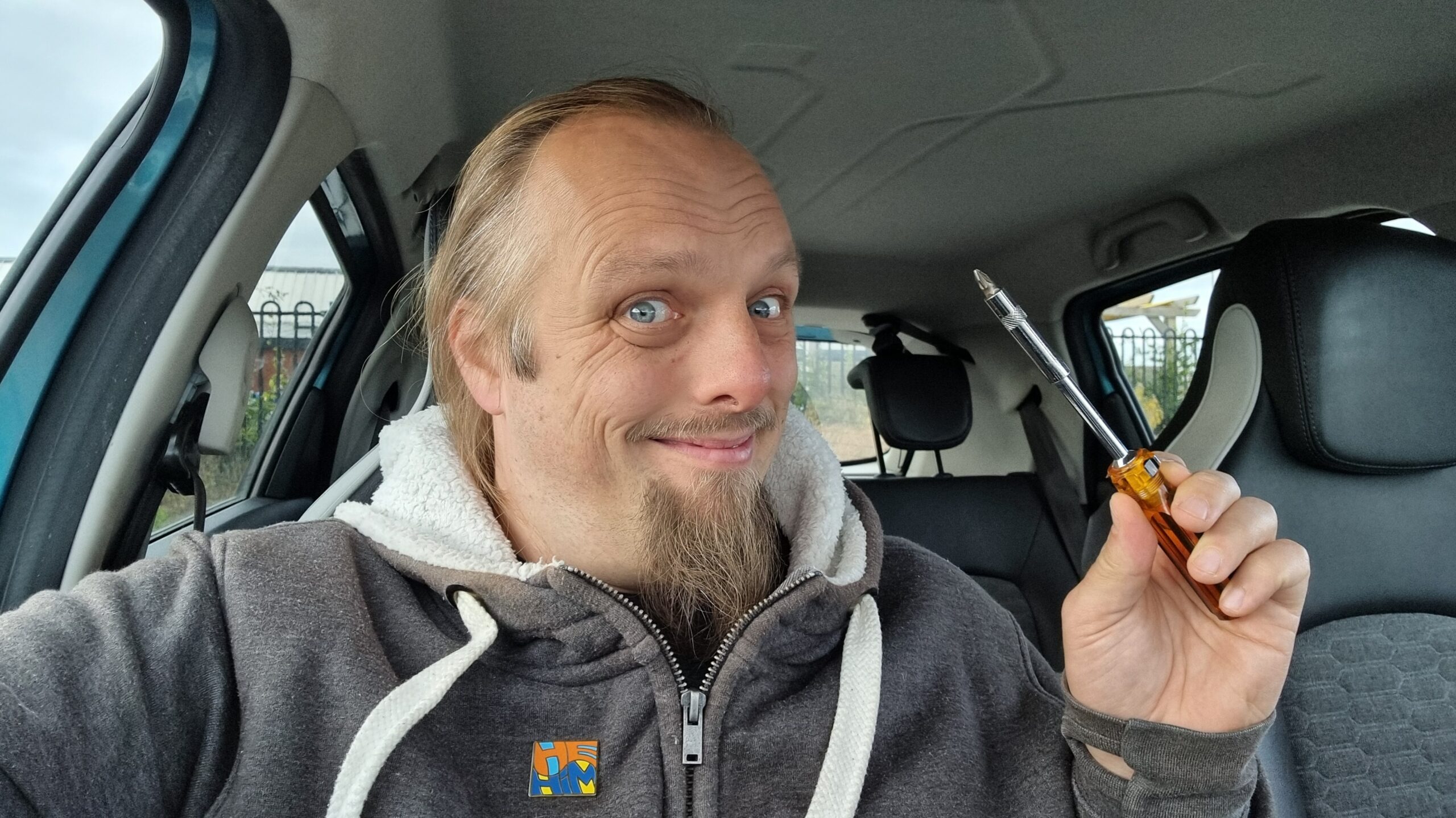 Full image of Dan, sitting in a car, holding a screwdriver and smiling.