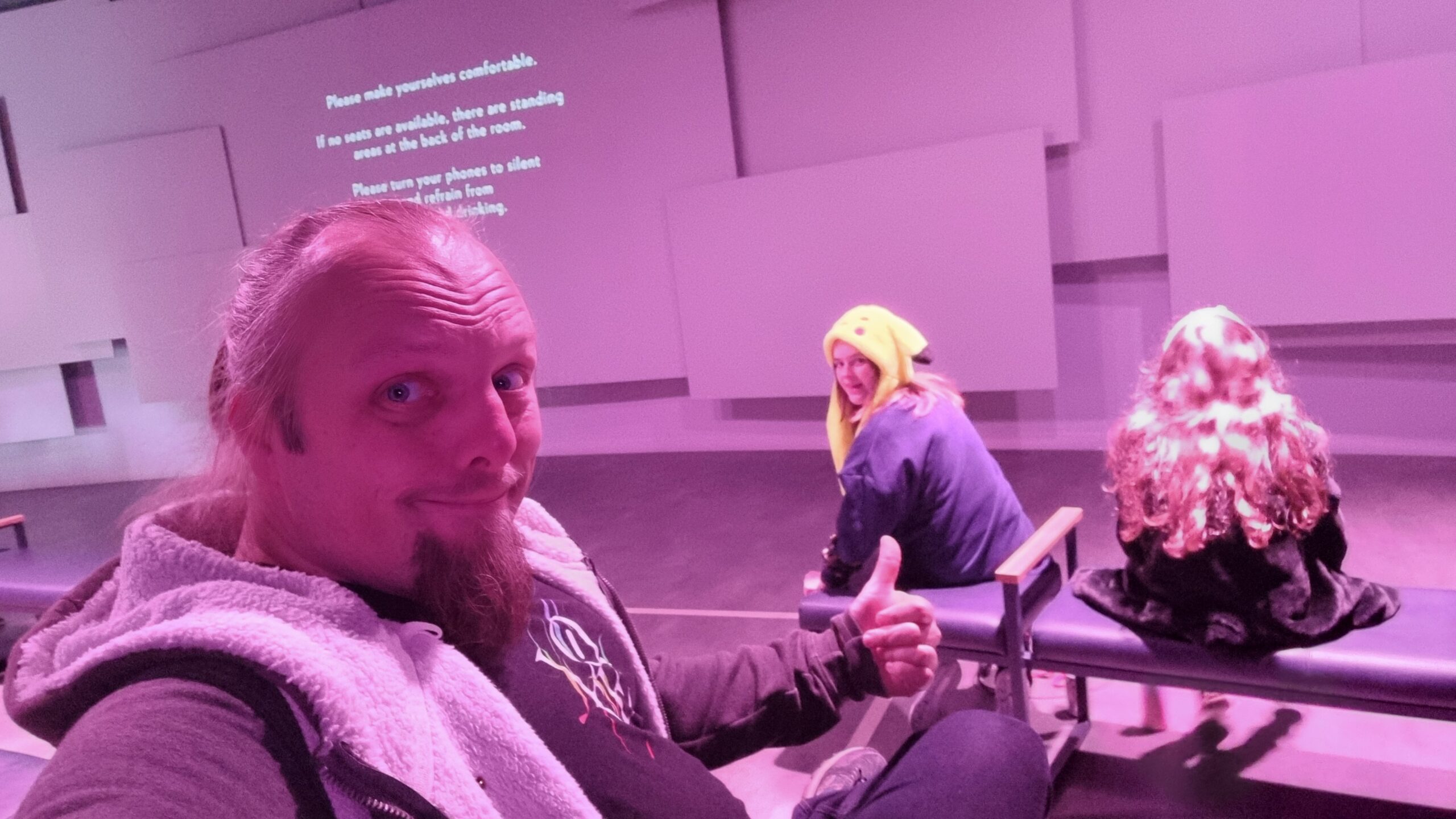 Full image of Dan sits on a padded bench in a cinema-style room, pointing his thumb at the two children sitting on a row in front of him.