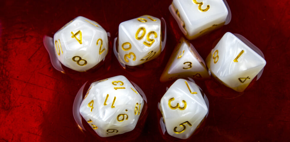 A 'full set' of white polyhedral dice commonly-used by roleplayers - a D4, D6, D8, two D10s, a D12, and a D20 - sit half-submerged in a red liquid.