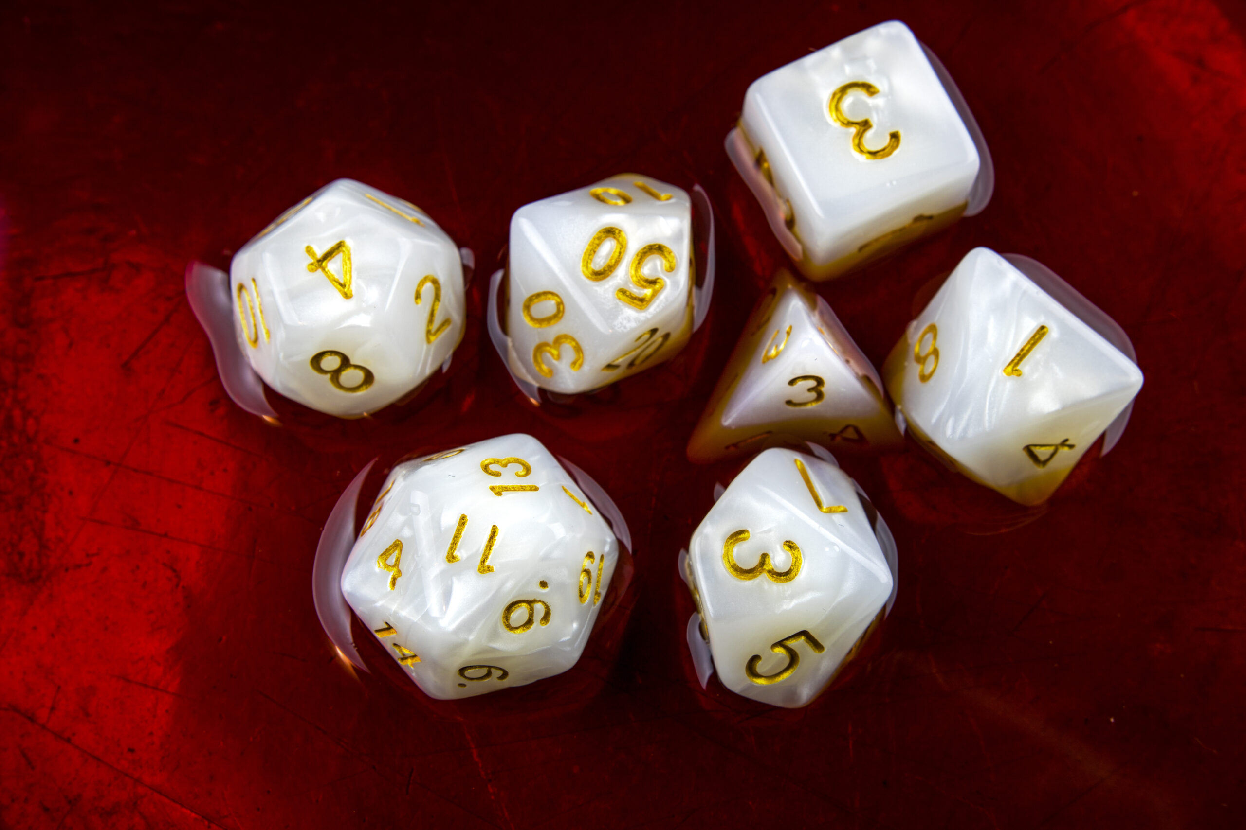 Full image of A 'full set' of white polyhedral dice commonly-used by roleplayers - a D4, D6, D8, two D10s, a D12, and a D20 - sit half-submerged in a red liquid.