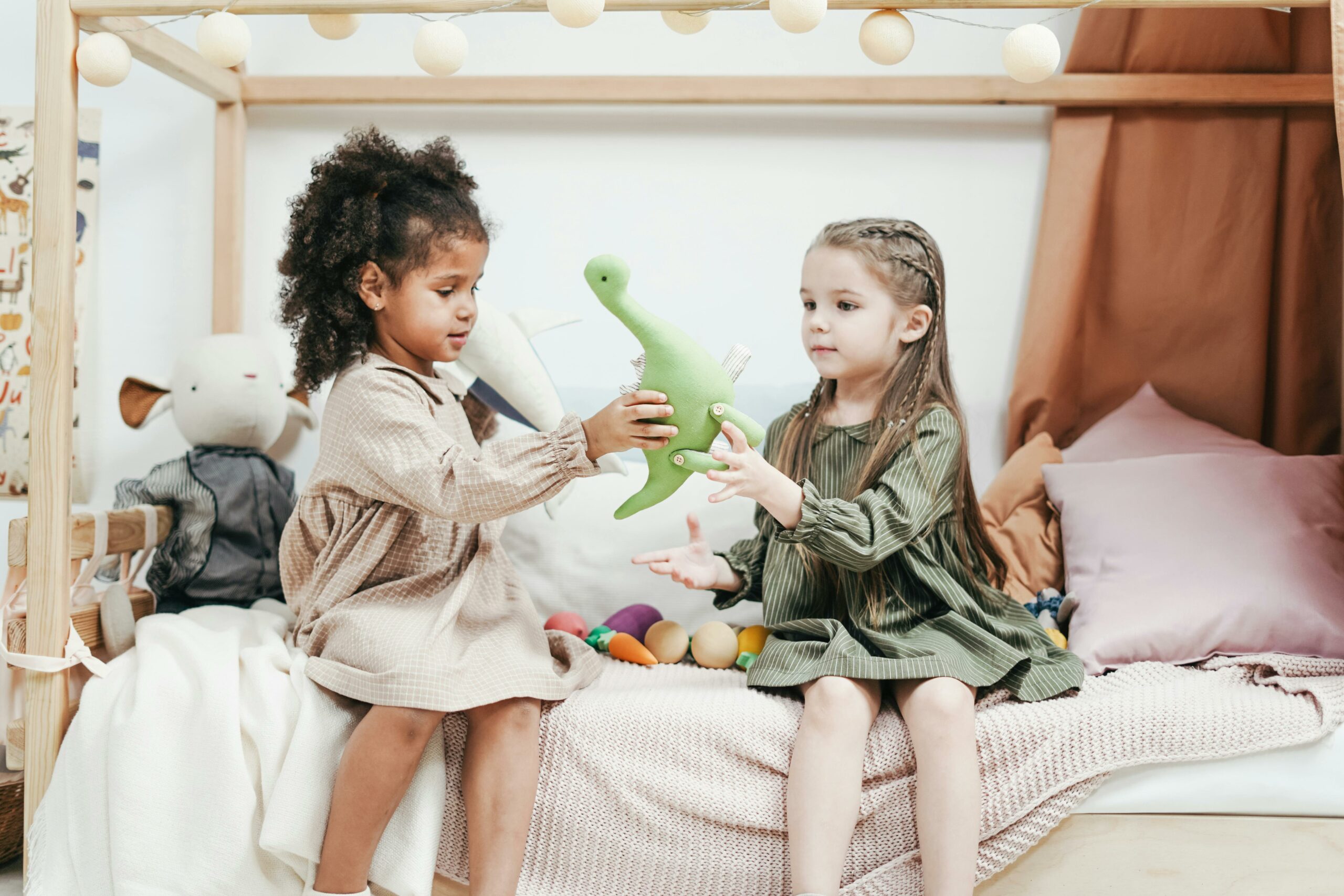 Full image of Two children sit on a bed: one hands a toy dinosaur to the other.