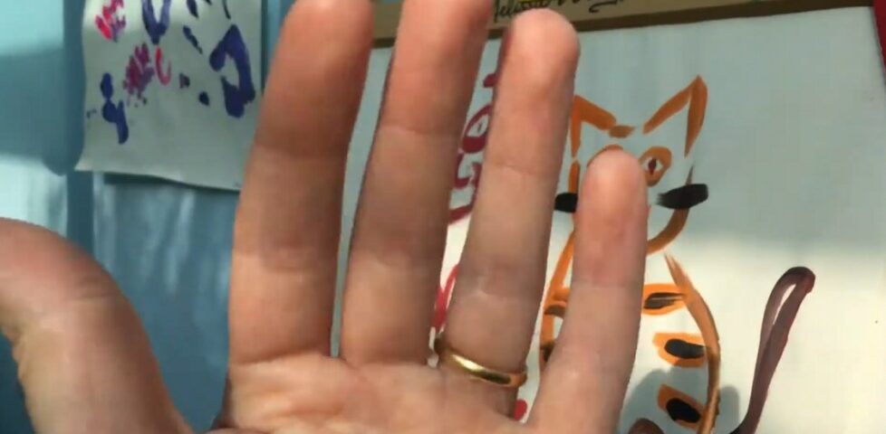 Screengrab showing the fingers of a white hand, one wearing a gold ring, in front of a child's(?) painting.