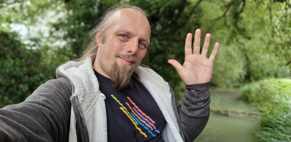 Dan, sitting on a bridge edge near a wooded stream, waves to the camera.