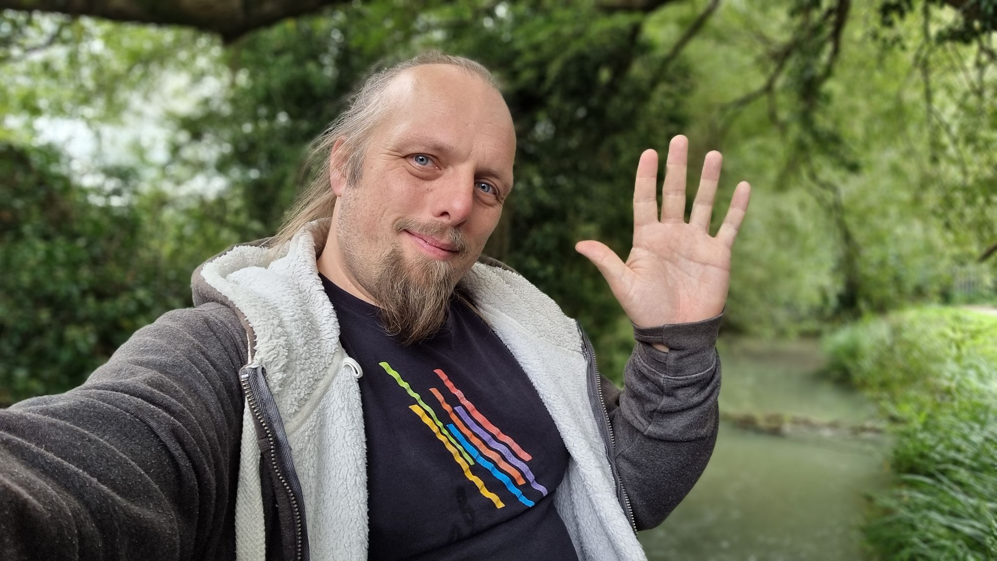 Full image of Dan, sitting on a bridge edge near a wooded stream, waves to the camera. 