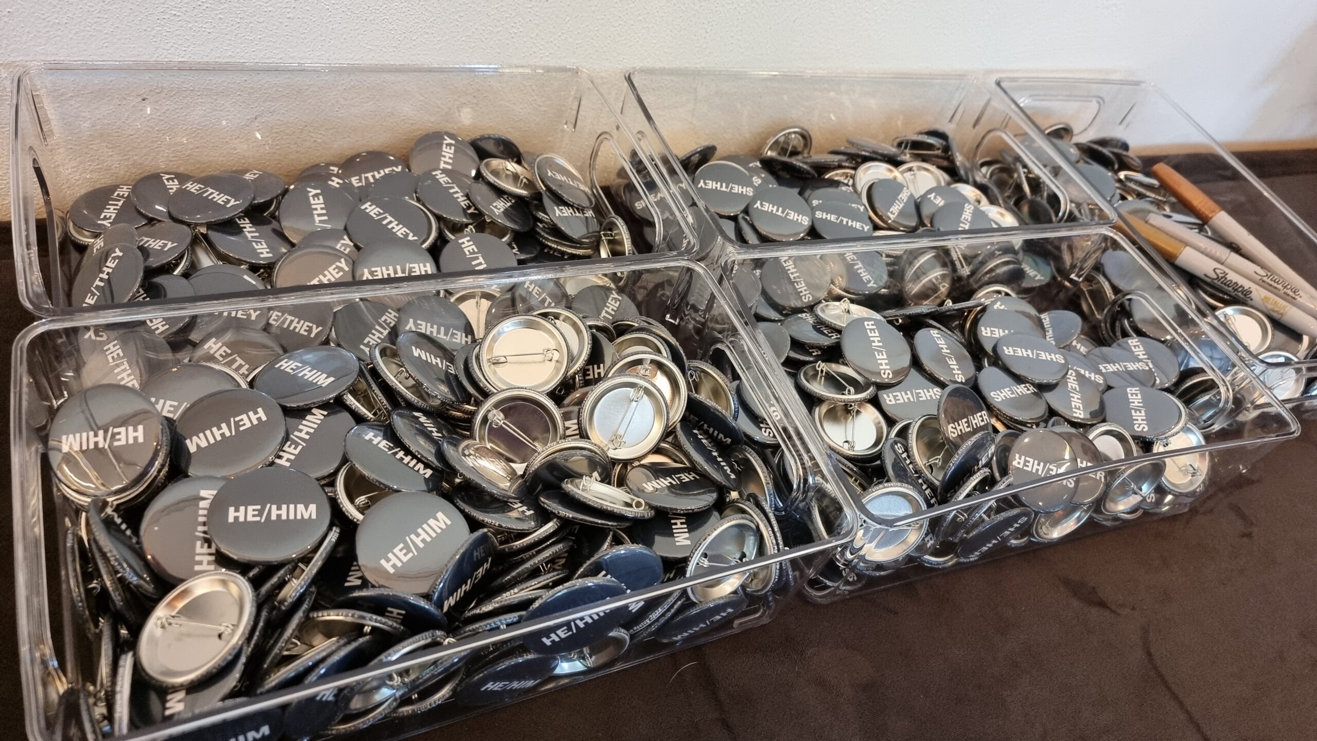 Full image of Boxes of pin badges representing various pronouns, plus blank ones and a sharpie.