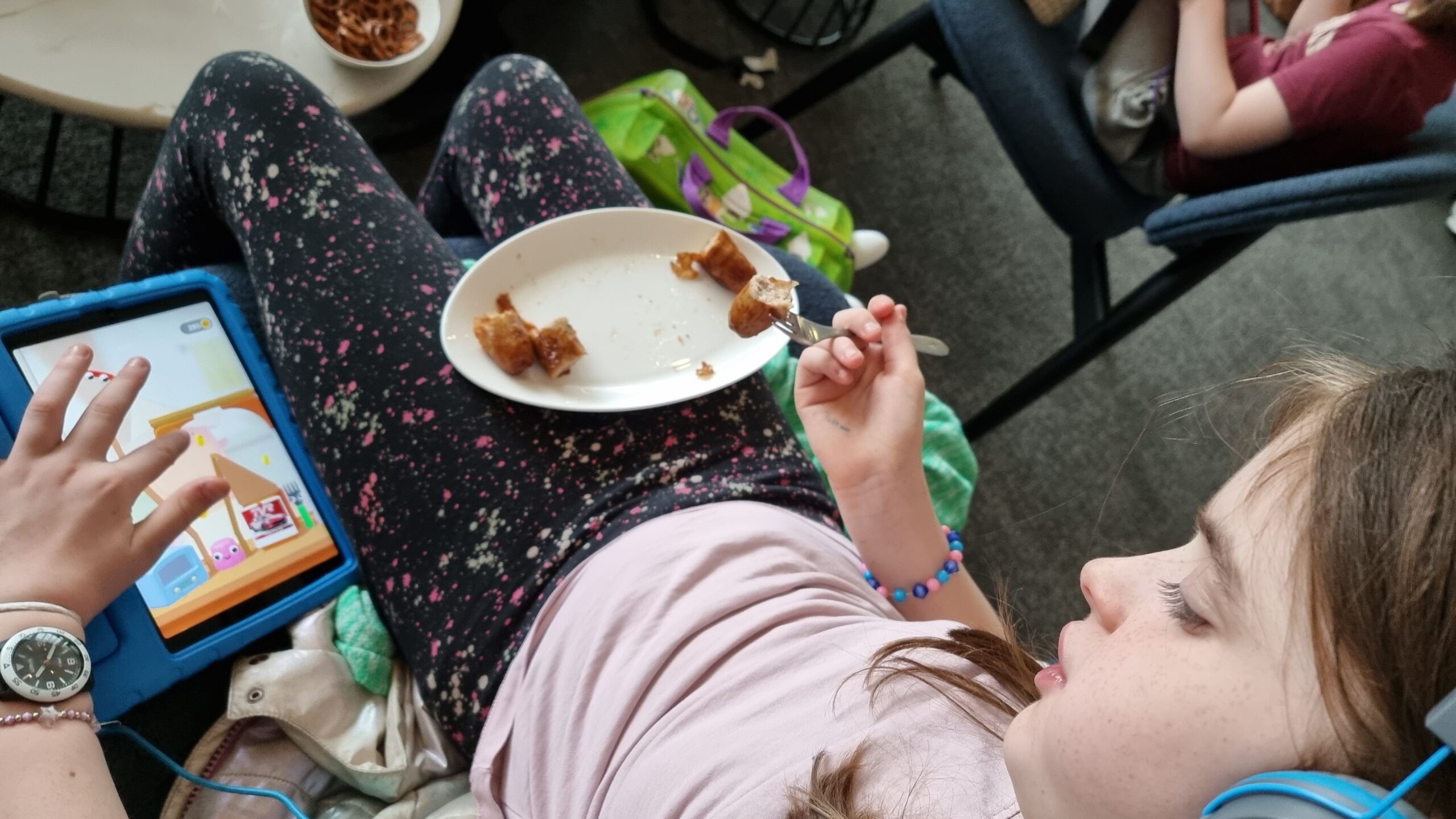Full image of A child slouches in an airport lounge chair. Her right hand is being used to eat a plate of sausages. Her left hand is playing 'Fork N Sausage' on a tablet.